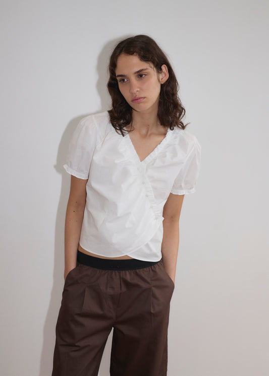 A person with wavy brown hair wears a white V-neck blouse and Deiji Studios Contrast Waist Pant - Brown, standing against a plain light wall with hands in pockets, looking slightly down and to the side.
