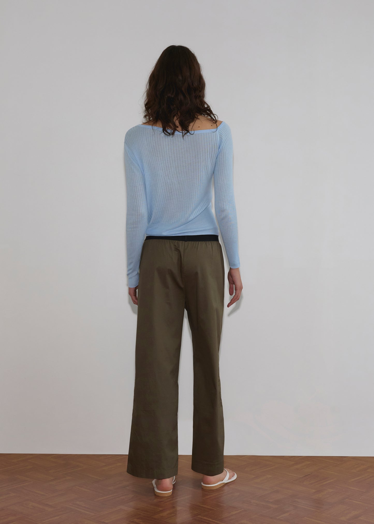 A person with wavy brown hair faces away from the camera, wearing a light blue Shoulder Knit Top - Blue from Deiji Studios, olive-green wide-leg pants, and pink shoes on a wooden floor against a plain white wall.