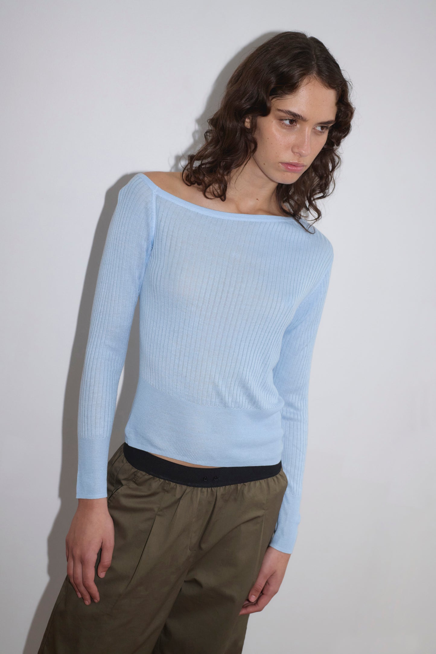 A person with wavy brown hair wears the Shoulder Knit Top - Blue from Deiji Studios with olive green pants, standing against a plain white background and looking slightly downward.