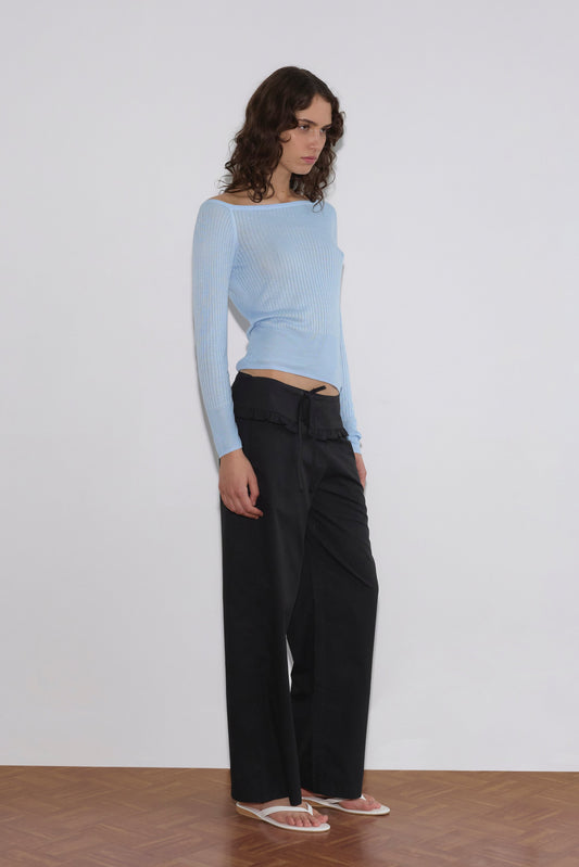 A person with wavy brown hair stands against a white wall on a wooden floor, wearing a light blue off-shoulder ribbed top, Flounce Belt Pant in black, and white flip-flops.
