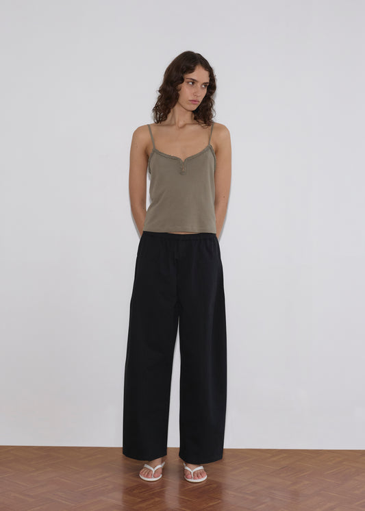 A woman with wavy brown hair stands against a plain white wall, wearing a khaki camisole, Deiji Studios Pleat Pant in Black, and white sandals. Her hands are behind her back as she gazes slightly to the side.