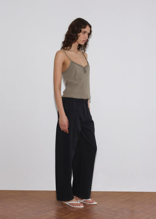 A person with long, wavy brown hair stands sideways against a plain white wall, wearing a taupe camisole, Pleat Pant - Black by Deiji Studios, and clear sandals on a wooden floor.
