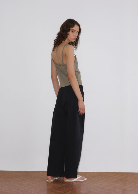 A person with long curly hair stands on a wooden floor, slightly turned toward the camera. They wear the Clasp Singlet - Khaki by Deiji Studios with adjustable straps, loose black pants, and white sandals against a plain white background.