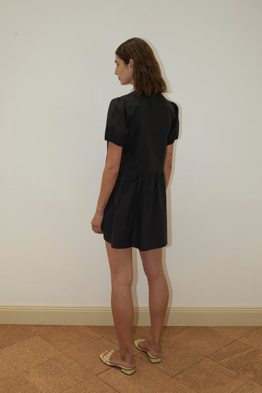 A person with shoulder-length brown hair stands sideways against a white wall and brown tiled floor, wearing the Pocket Mini Dress - Black by Deiji Studios with short sleeves and beige sandals.