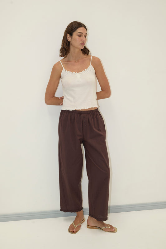 A woman with wavy brown hair stands against a plain white wall wearing the Wool Blend Flounce Tank - Cream, loose brown pants, and clear sandals, with her hands in her pockets.