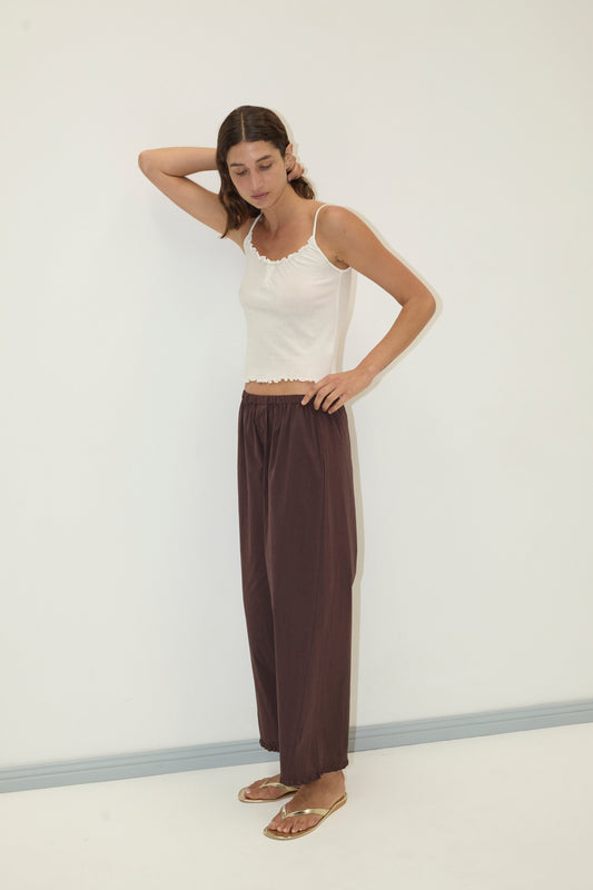 A woman stands against a white wall wearing the Deiji Studios Wool Blend Flounce Tank in cream, loose brown pants, and gold sandals. One hand is behind her head as she looks slightly downward.