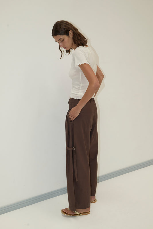 A person with brown hair wears a fitted white short-sleeve top, Tab Pant - Dark Brown, and flat sandals, standing sideways against a plain light-colored wall and floor.