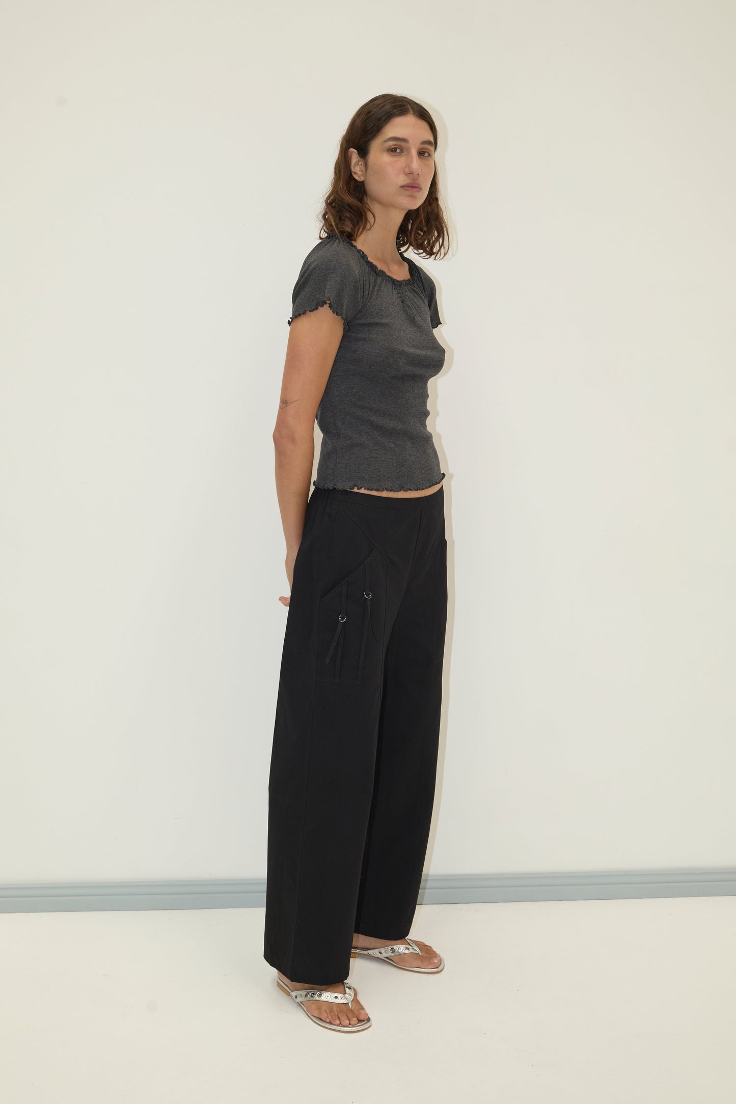 A woman with wavy brown hair stands against a plain light wall, wearing the Flounce Tee - Grey by Deiji Studios, wide-leg black pants with pockets, and sandals. She looks slightly to the side.