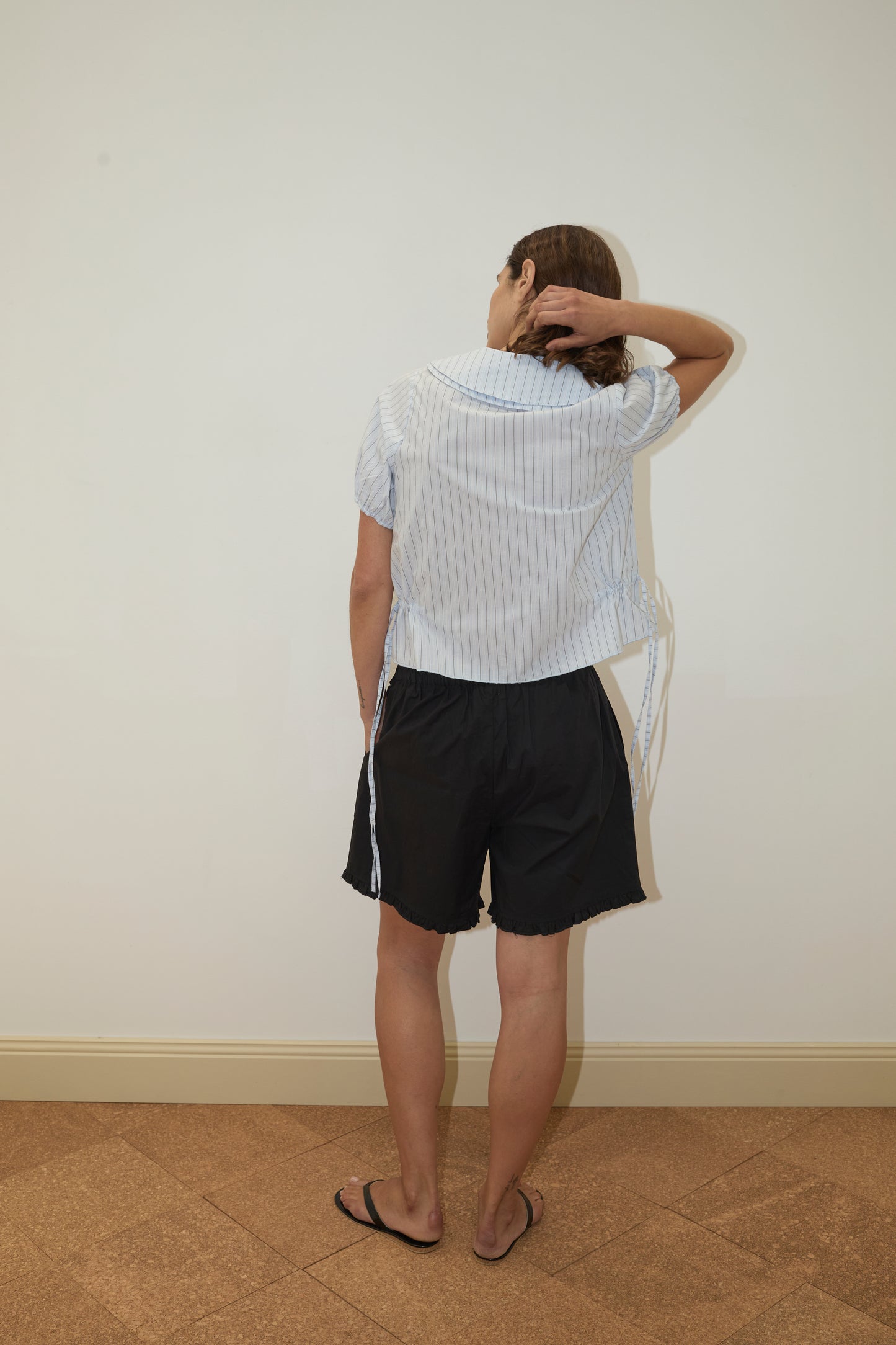 A person faces a blank wall with their back to the camera, hand on head, wearing the Deiji Studios Double Collar Shirt - Mineral Stripe, paired with black shorts and black flip-flops.