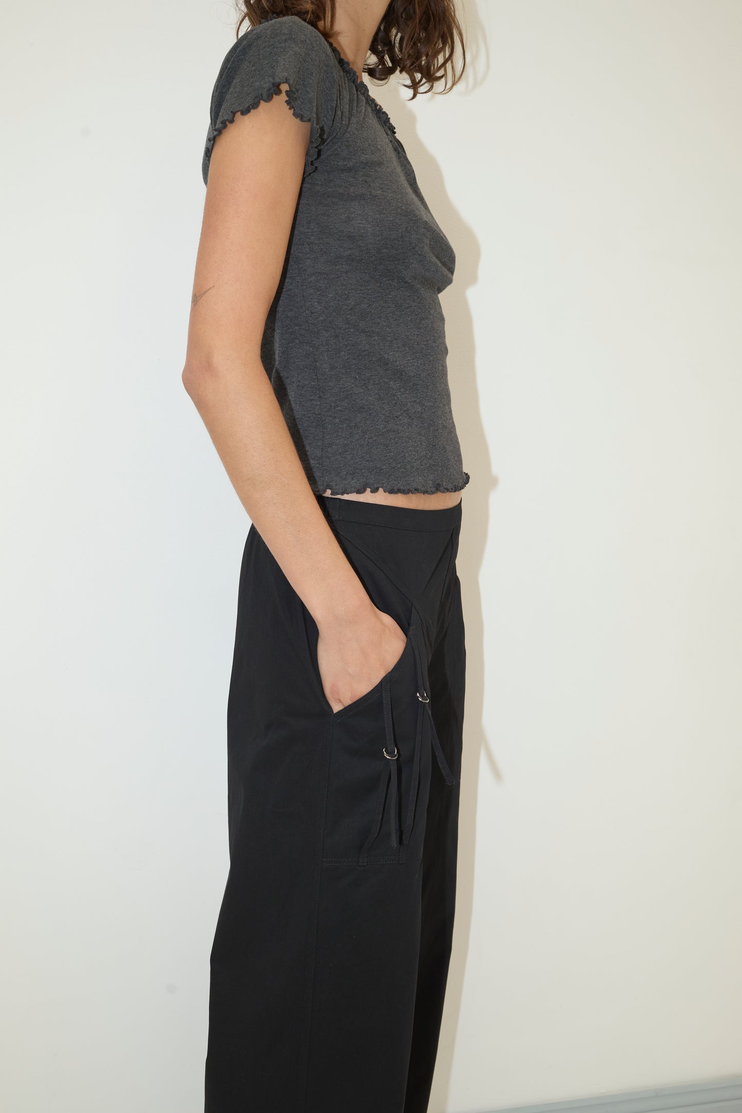 A woman stands sideways against a plain white wall, wearing a fitted gray short-sleeve top and the Adjust Pocket Pant - Black by Deiji Studios in loose organic cotton, her hand tucked in the pocket. Her head is not visible in the image.