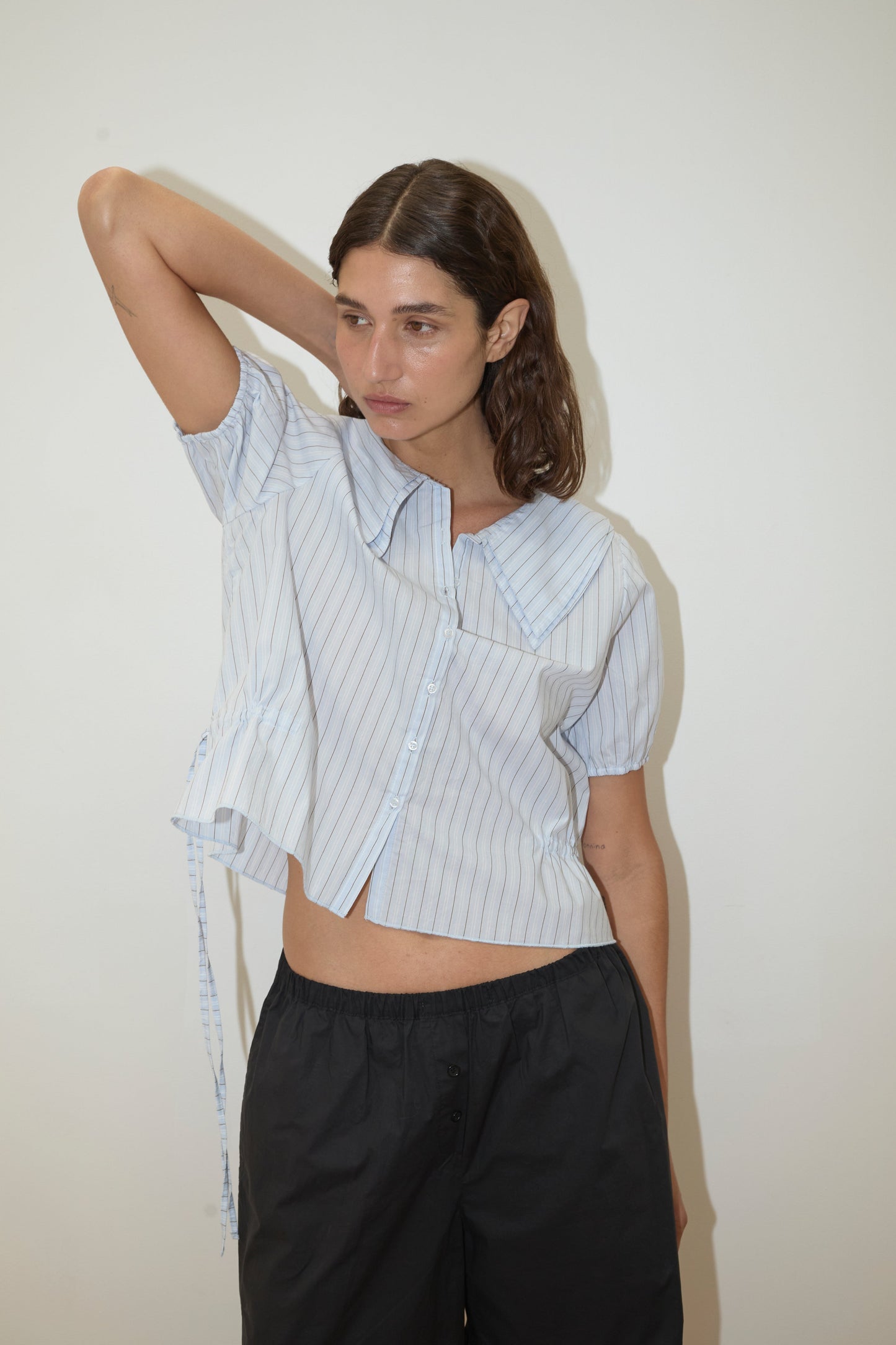 A woman with brown hair poses against a white background, wearing the Double Collar Shirt - Mineral Stripe by Deiji Studios and loose black pants. She raises one arm behind her head.