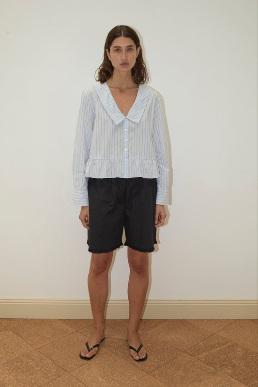 A person stands indoors against a plain white wall, wearing a light blue striped blouse, Flounce Short - Black with an elastic waist, and black flip-flops.