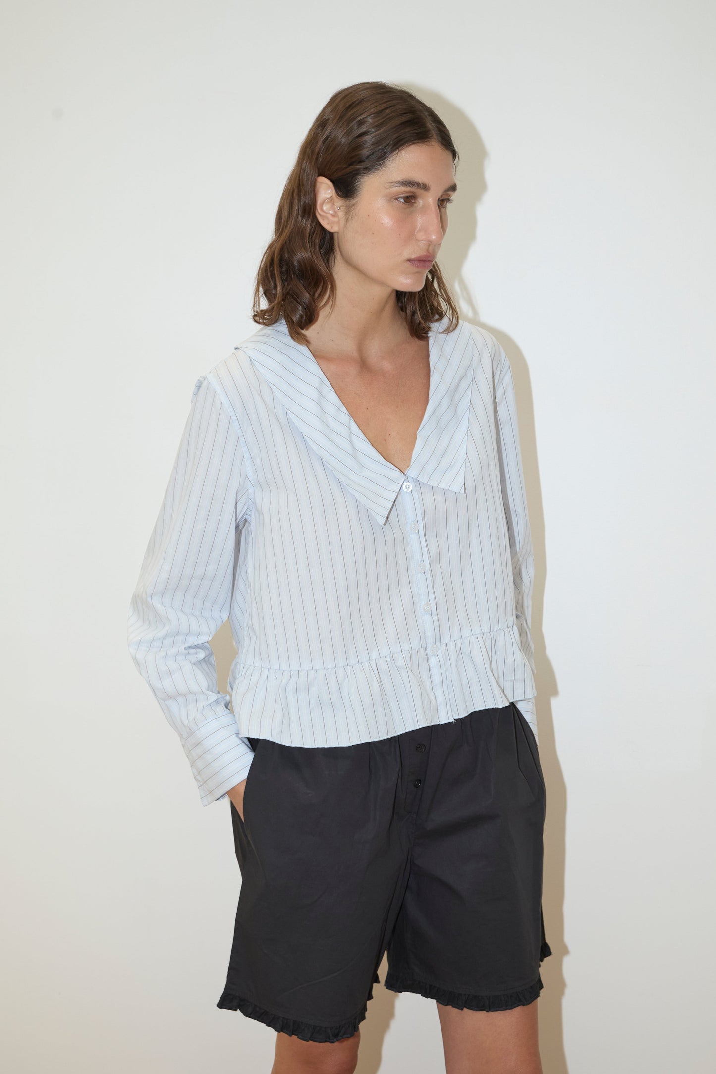 A woman with wavy brown hair stands against a plain white background, wearing the Deiji Studios Sweep Shirt - Mineral Stripe with light blue and white stripes and black shorts. She has one hand in her pocket and gazes slightly to the side.