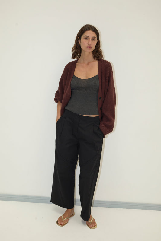 A woman stands against a white wall wearing the Flounce Cardigan - Burgundy over a gray ribbed tank top, loose black pants, and gold sandals, with her hands in her pockets and a neutral expression.