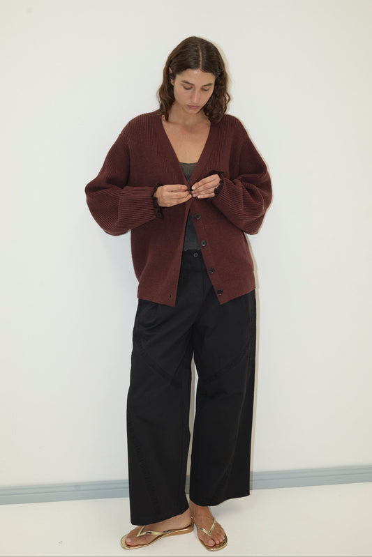 A woman stands against a white wall, looking down as she buttons the Flounce Cardigan - Burgundy by Deiji Studios over a dark top, paired with wide-leg black pants and gold sandals.