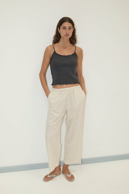 A woman with long brown hair stands against a white wall, wearing the Wool Blend Flounce Tank - Grey by Deiji Studios, loose light beige pants, and flip-flops. Her hands rest casually in her pockets.