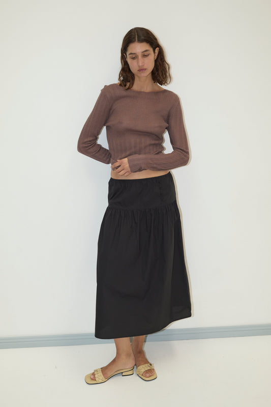 A woman stands against a plain white wall, wearing the Open Work Longsleeve in Hazel, a black ankle-length skirt, and beige sandals. She looks down with her hands resting on her skirt.