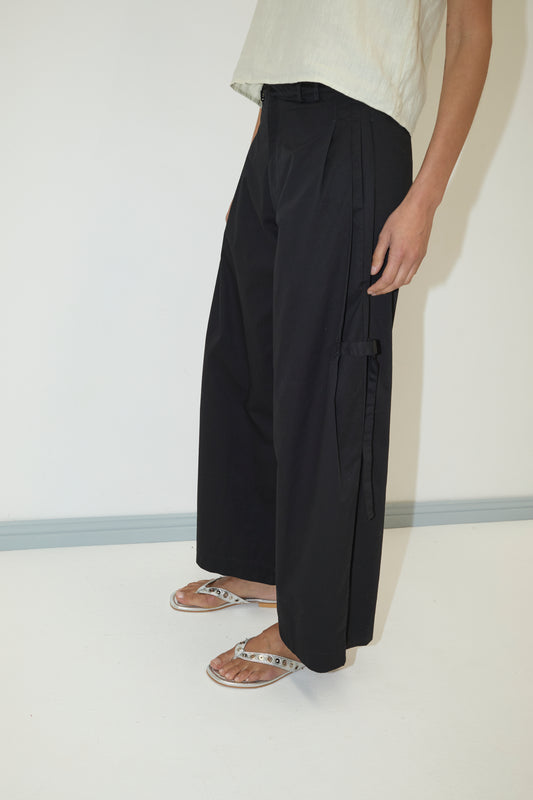 Only the lower half of a person is visible as they stand against a white wall, wearing a light top, Tab Pant - Black by Deiji Studios with side pockets, and silver flip-flop sandals.