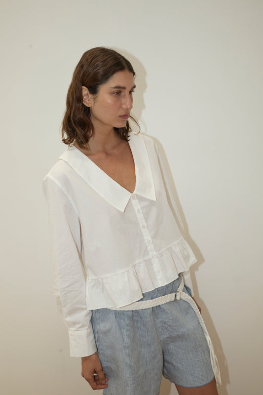A person with wavy brown hair wears the Deiji Studios Sweep Shirt - White, a loose button-up blouse, paired with light blue and white striped shorts, standing against a plain off-white wall.