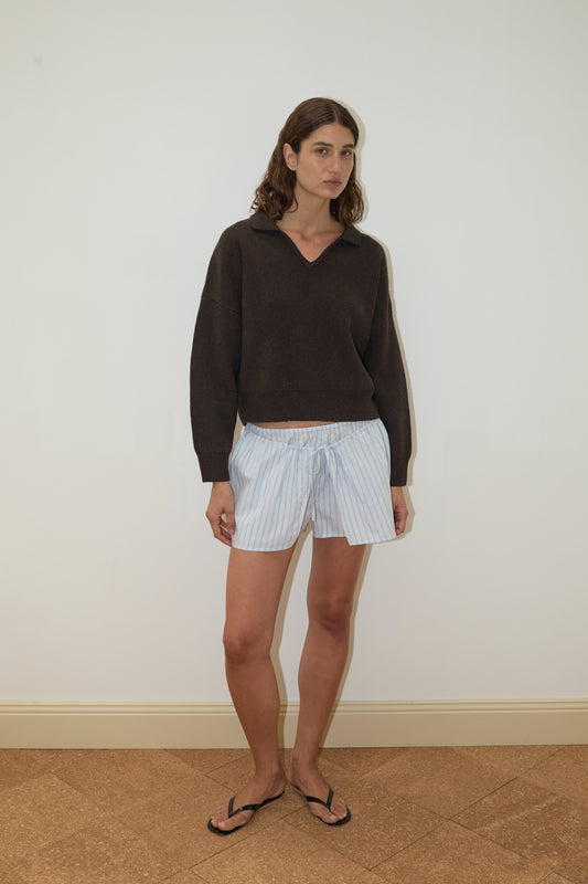 A woman stands indoors against a plain white wall, wearing the Deiji Studios Collared Pullover - Burnt Wood, light blue and white striped shorts, and black flip-flops. She has wavy brown hair and a neutral expression.