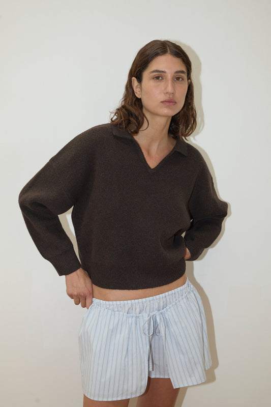 A woman with wavy brown hair stands against a plain white background, wearing the Deiji Studios Collared Pullover in Burnt Wood and light blue, drawstring, striped shorts. She looks at the camera with a neutral expression and one hand on her hip.
