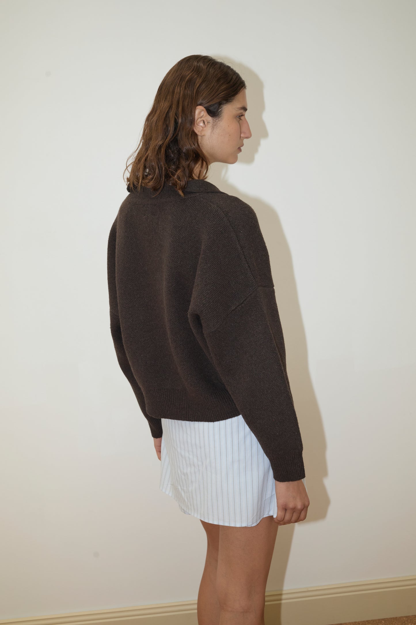 A person with wavy brown hair stands sideways, wearing the Deiji Studios Collared Pullover in Burnt Wood and a short, light blue pinstriped skirt, against a plain light-colored wall.