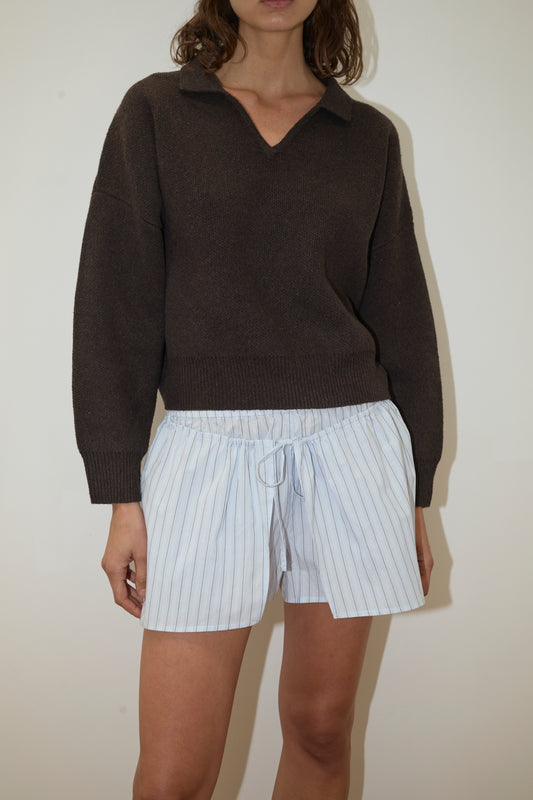 A person stands against a plain background, wearing the Collared Pullover - Burnt Wood by Deiji Studios and loose white drawstring shorts with thin blue stripes. The person's face is not fully visible.