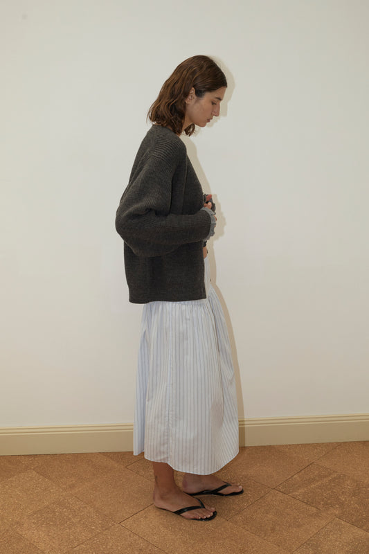 A person with shoulder-length brown hair stands in profile against a plain wall, wearing a Deiji Studios Flounce Cardigan - Charcoal, a long white pleated skirt with subtle stripes, and black sandals on a light brown patterned floor.