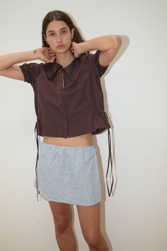 A person with long brown hair stands against a plain white wall, wearing the Double Collar Shirt—a short-sleeved dark brown button-up top with ties—and a light blue mini skirt, arms raised to adjust their collar and face neutral.