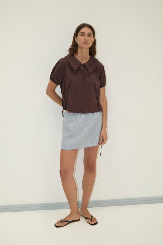 A woman with long brown hair and a neutral expression stands against a white wall wearing the Deiji Studios Double Collar Shirt - Umber with blouson sleeves, a light blue patterned mini skirt, and black flip-flops.