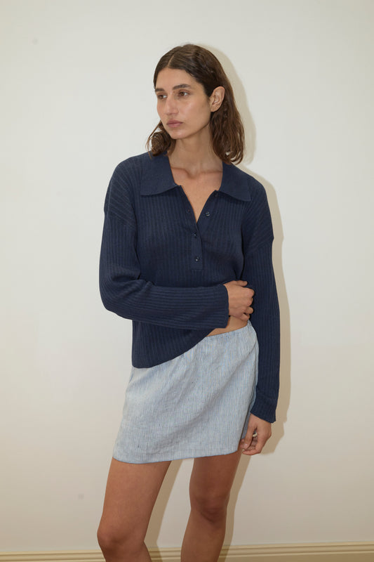 A woman with shoulder-length brown hair wears the Polo Knit - Dark Blue top, paired with a light blue mini skirt, standing against an off-white wall and looking to the side.