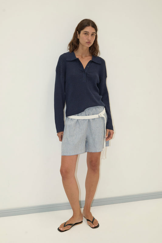 A woman with wavy brown hair stands against a white wall in a relaxed pose, wearing a Deiji Studios Polo Knit - Dark Blue, light blue patterned shorts with a white belt, and black flip-flops.