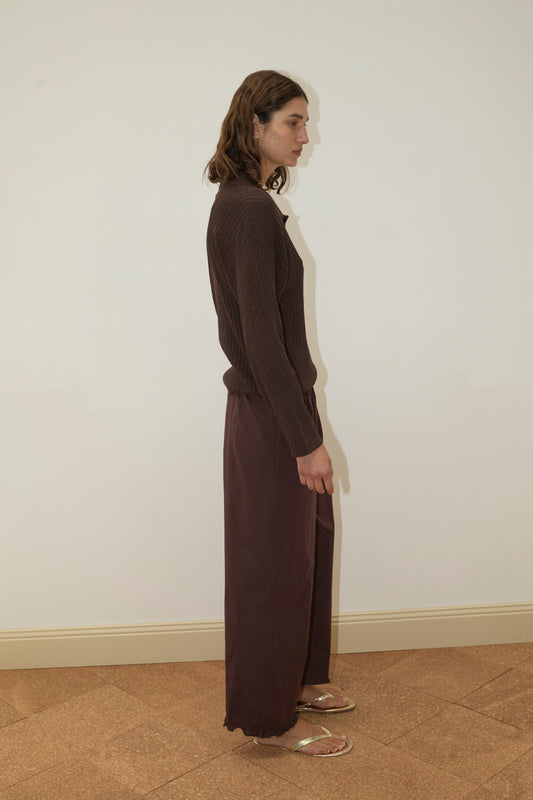 A person with shoulder-length brown hair, wearing the Deiji Studios Polo Knit - Dark Almond long sleeve top, wide-legged brown pants, and sandals, stands in profile against a plain off-white wall and tan floor.