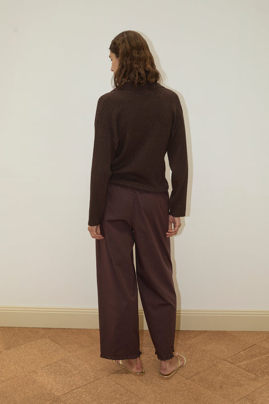 A person with shoulder-length brown hair stands barefoot, facing a plain off-white wall with their back to the camera, wearing the Deiji Studios Polo Knit - Dark Almond and loose-fitting brown pants.