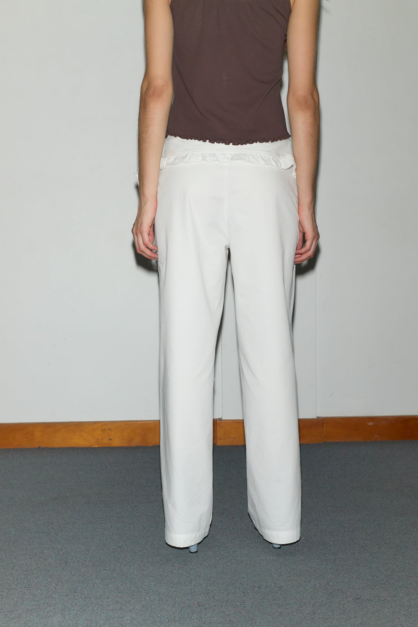 A person stands facing away from the camera, wearing a sleeveless brown top and the Flounce Belt Pant - White against a plain wall with wooden trim and a gray carpeted floor.