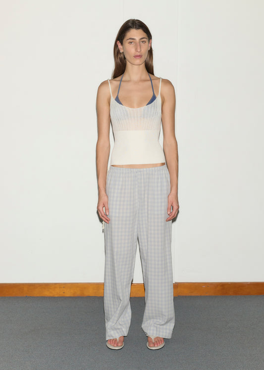 A person stands indoors against a plain white wall, wearing a cream fitted tank over a blue bralette, Side Tie Pant - Blue Check, and white flip-flops. Their expression is neutral with arms relaxed at their sides.