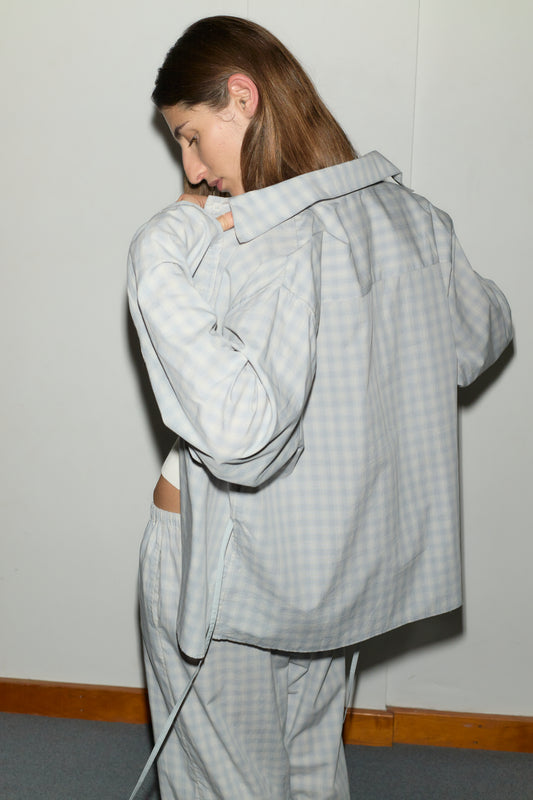 A person with long brown hair wearing the Day Shirt - Blue Check, a boxy fit, light-colored check pajama top, looks down and gently pulls the collar to reveal part of their back against a plain light wall.