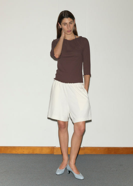 A woman stands indoors against a white wall, wearing a brown Cotton Flounce 3/4 Sleeve top by Deiji Studios, loose white shorts, and light blue heels. She touches her neck with one hand and gazes downward with a neutral expression.