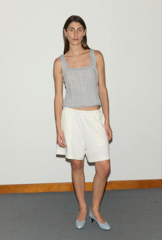 A model wears a Deiji Studios Button Frill Top in Blue Check, cream knee-length elastic waist shorts, and pale blue low-heel pumps, standing against a white wall.
