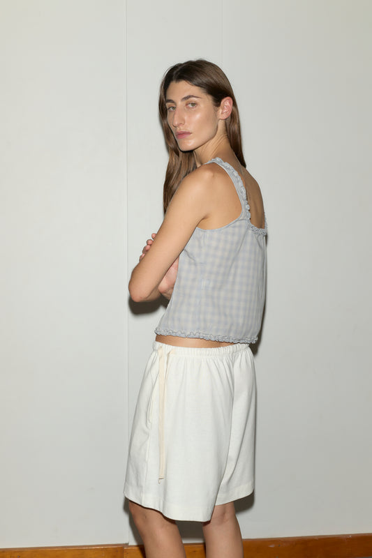 A woman with long brown hair stands against a plain light wall, wearing the Button Frill Top - Blue Check from Deiji Studios and loose white shorts, looking over her shoulder with arms crossed.