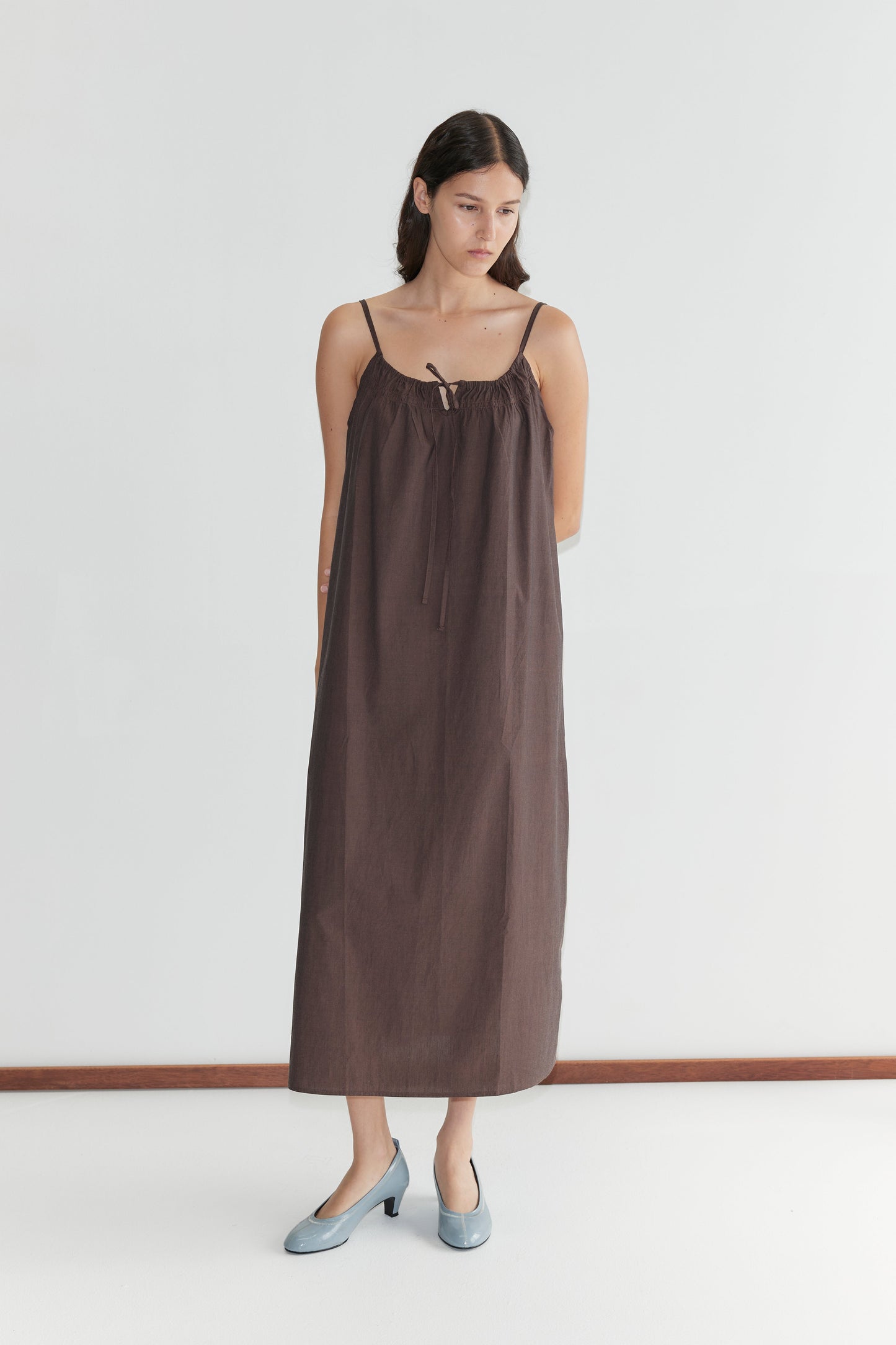 A woman with long dark hair wears the Light Maxi Dress - Toast by Deiji Studios, featuring thin straps and a relaxed fit, paired with light blue heels as she stands against a plain white background, gazing slightly to the side.