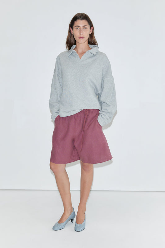 A person wearing a light gray sweatshirt, the "Half Short - Brick" linen shorts with an elasticated waistband and wide leg opening in a knee-length style, and blue heels stands against a plain white background, facing the camera with hands in their pockets.