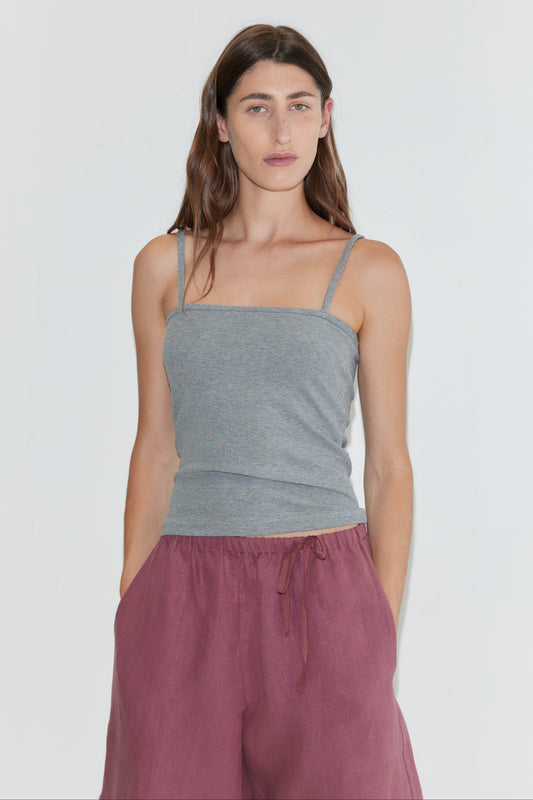 A woman with long brown hair, dressed in a gray spaghetti strap top and the Half Short - Brick, which features a wide leg opening and an elasticated waistband, stands against a plain white background with her hands in her pockets.