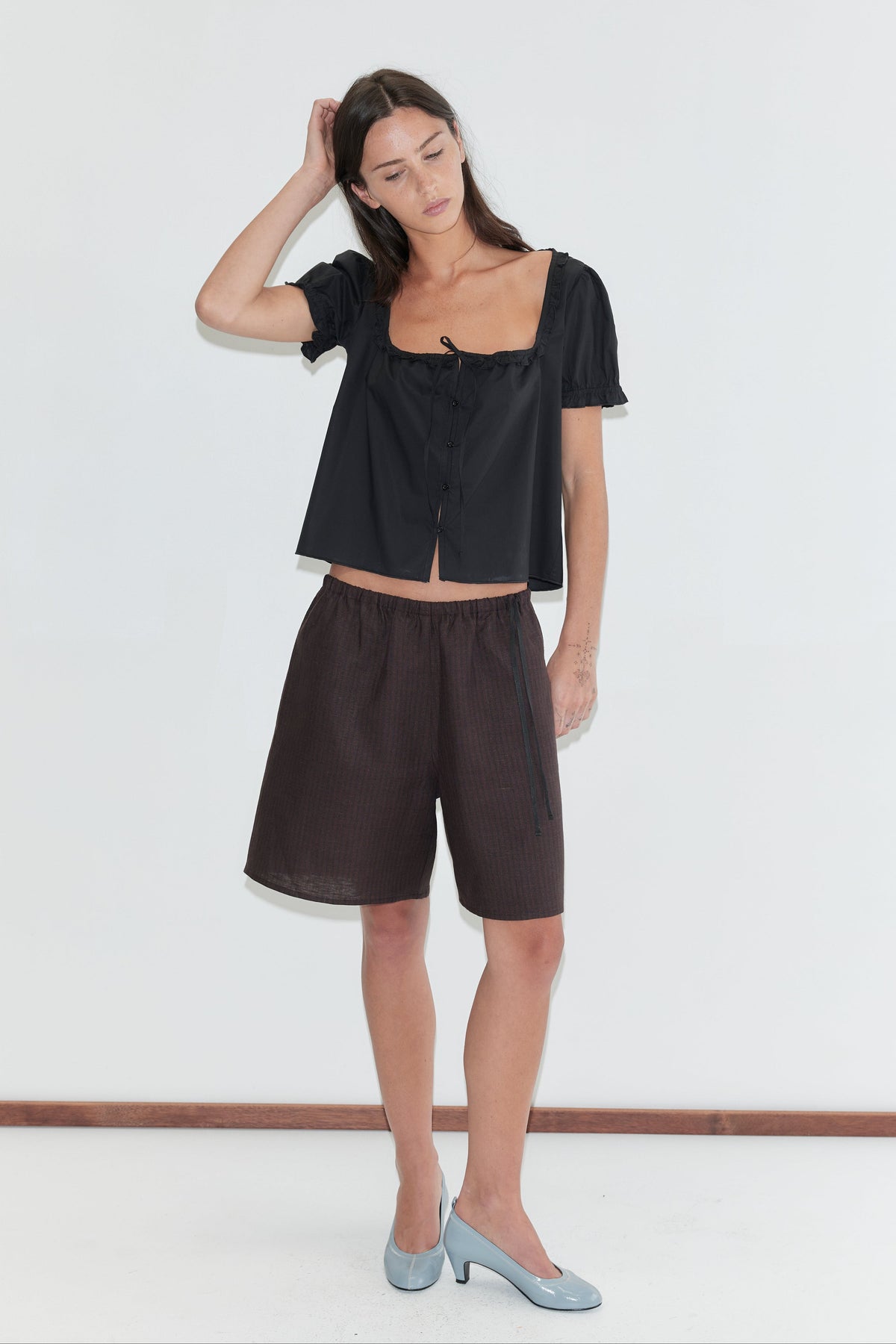 A woman with long dark hair stands against a plain white background wearing the Half Short - Brown Black Stripe by Deiji Studios, knee-length dark brown striped linen shorts, with a black flounced top and pale blue heels.