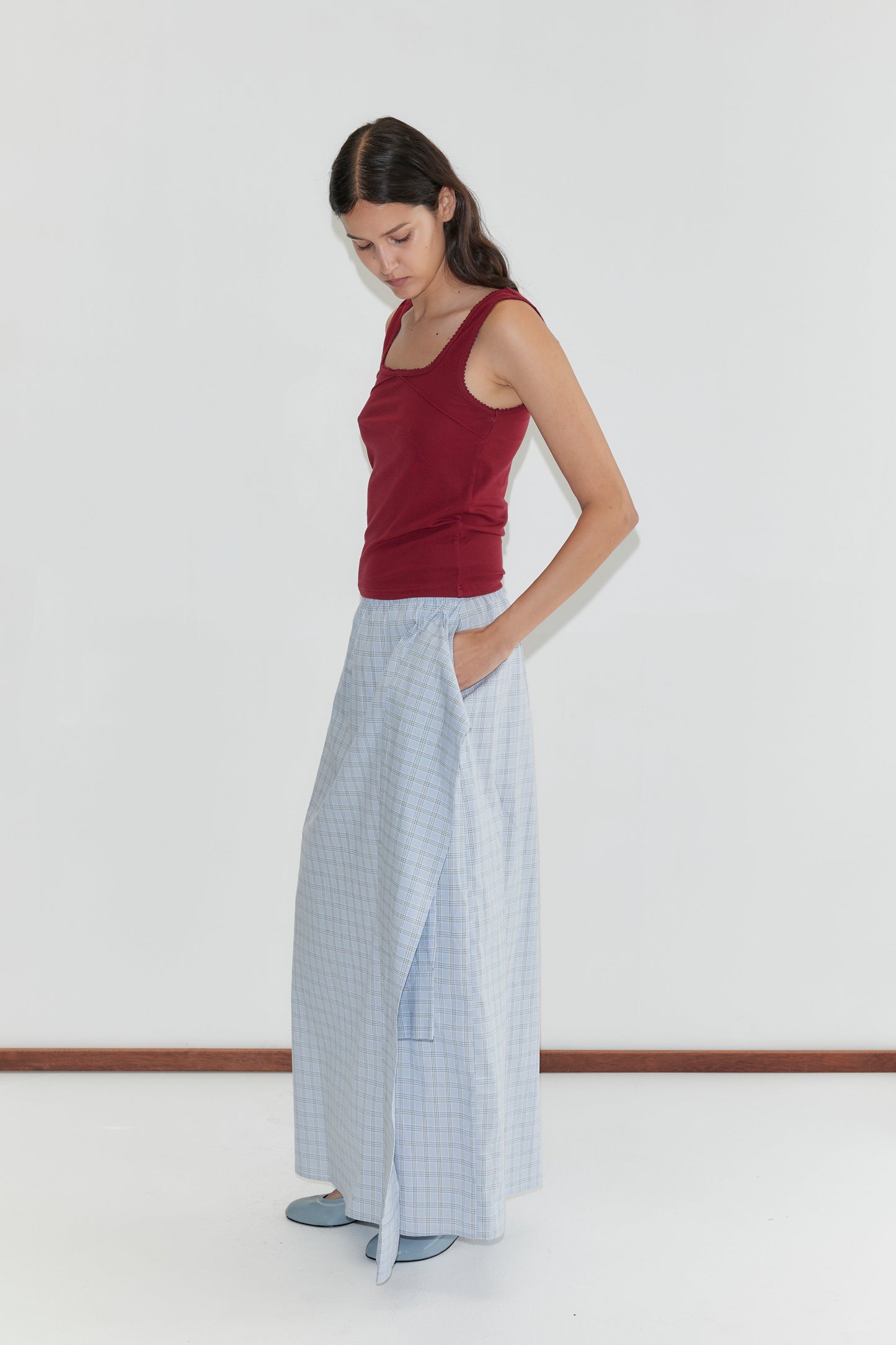 A woman stands against a white background, looking down. She wears a sleeveless maroon top, light blue checkered wide-leg pants with the Shirt Cuff Belt - Powder Check from Deiji Studios, blue shoes, and has long dark hair down with one hand in her pocket.