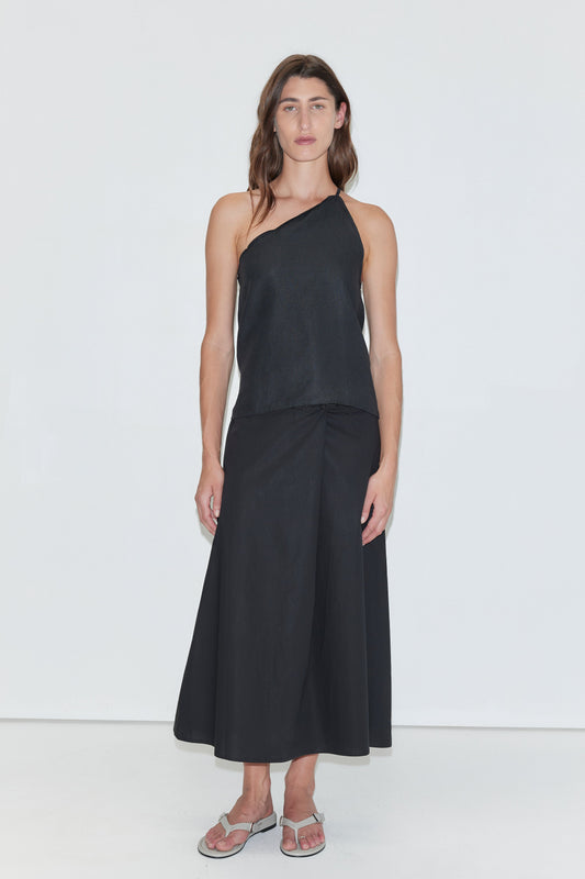 A person stands against a plain white background, wearing the Deiji Studios Half Halter - Black linen one-shoulder top, a long black skirt, and silver sandals. They have long brown hair and a neutral expression.