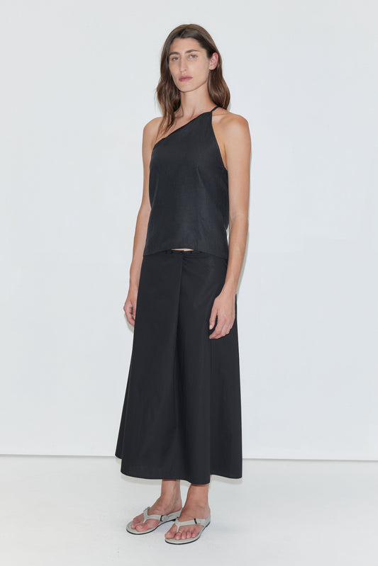 A woman stands against a plain white background wearing the Deiji Studios Half Halter - Black, paired with a black wide-leg skirt and silver flip-flops. She has long brown hair and a relaxed, neutral expression.