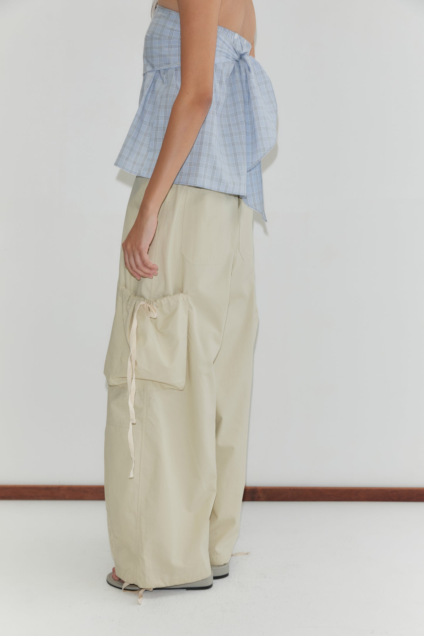 A person stands sideways against a white background, wearing a blue plaid strapless top with a large bow and high-waisted Deiji Studios Cargo Pant - Stone, featuring wide legs and an oversized pocket with ribbon tie.
