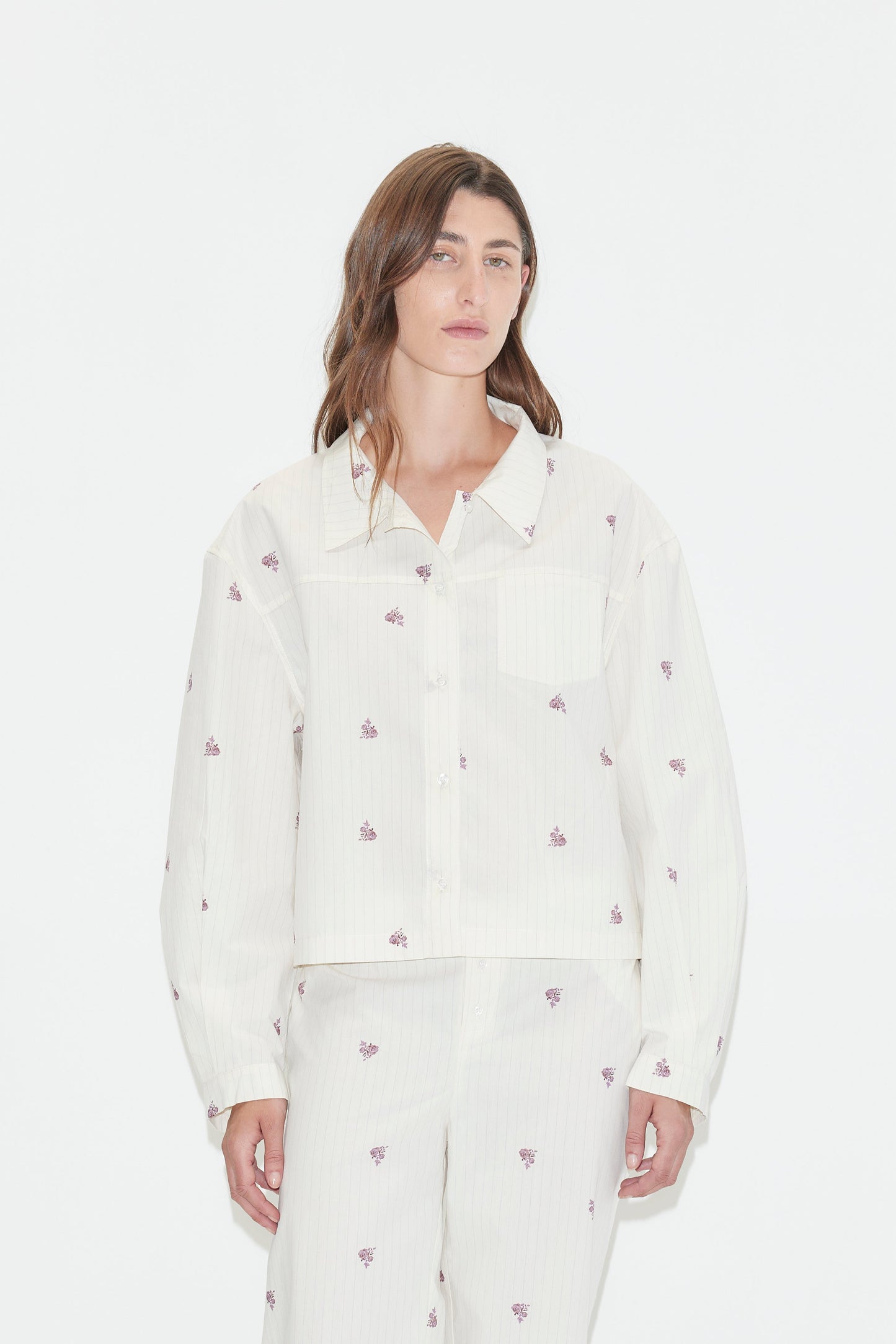 A person with long brown hair wears a loose-fitting white Boxy Shirt - Floral Stripe pajama set, featuring an oversized shirt adorned with small purple floral stripes, standing against a plain white background.