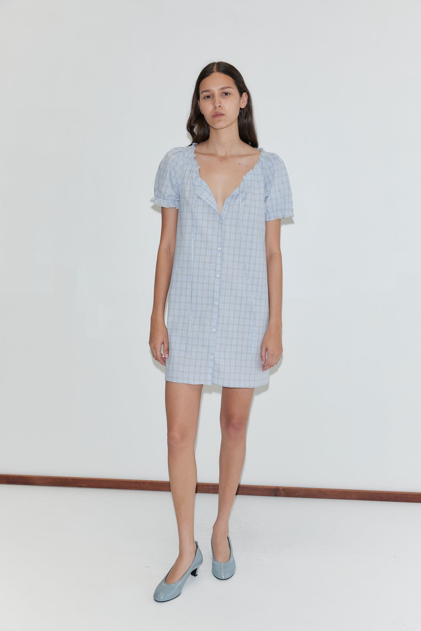 A woman with long dark hair wears the Buttoned Mini Dress - Powder Check and light blue heels, standing against a plain white background.